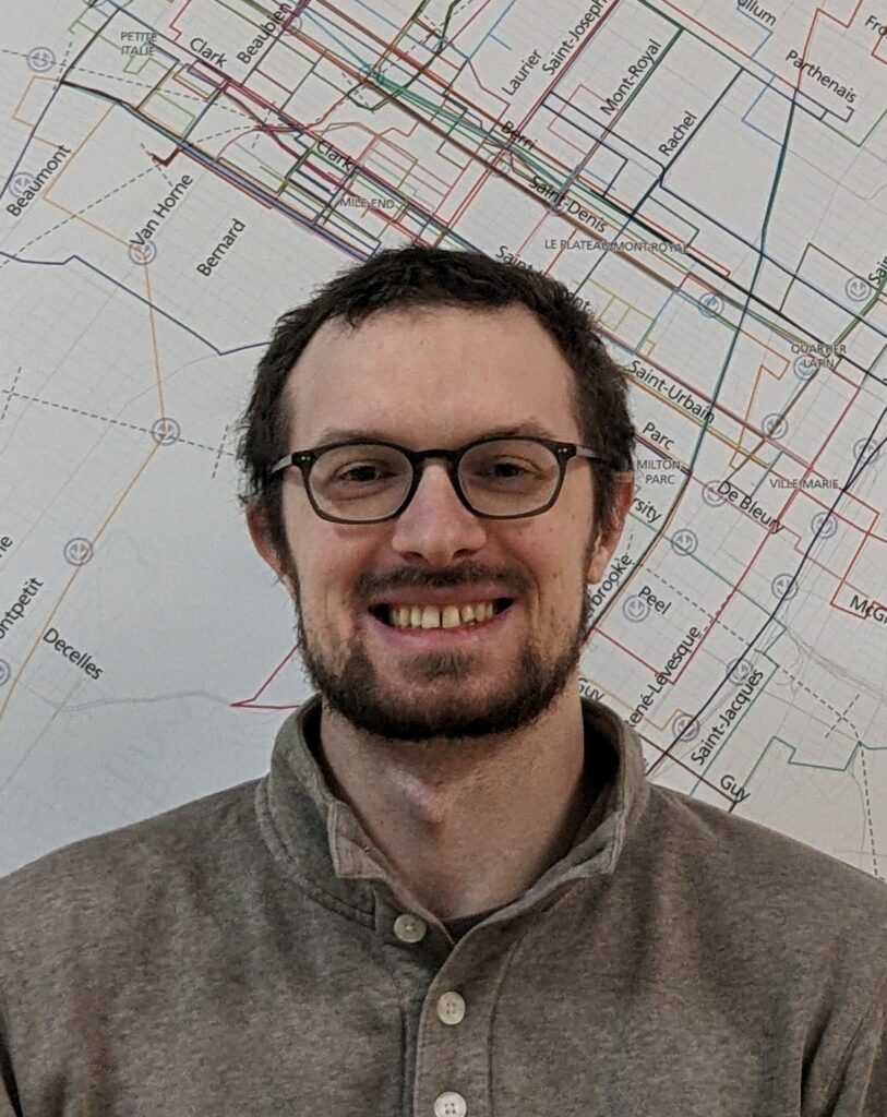 Colour photo of a smiling bearded man with glasses, standing in front of a large street map